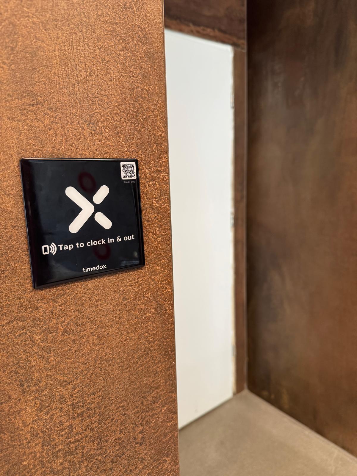 Timedox NFC tap-to-clock sticker mounted on a wall beside a doorway